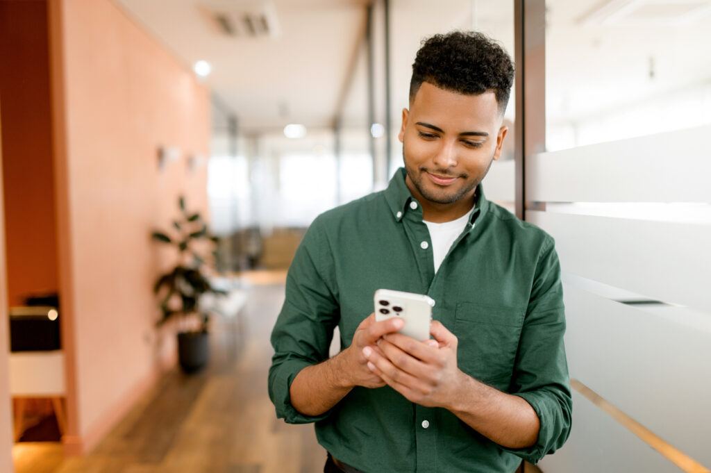 A real estate agent in a green shirt smiles while using his smartphone, standing in a modern office. This image illustrates the convenience of centralizing client communications on platforms like CryptoPay, enabling real estate agents to efficiently manage contacts and streamline interactions.