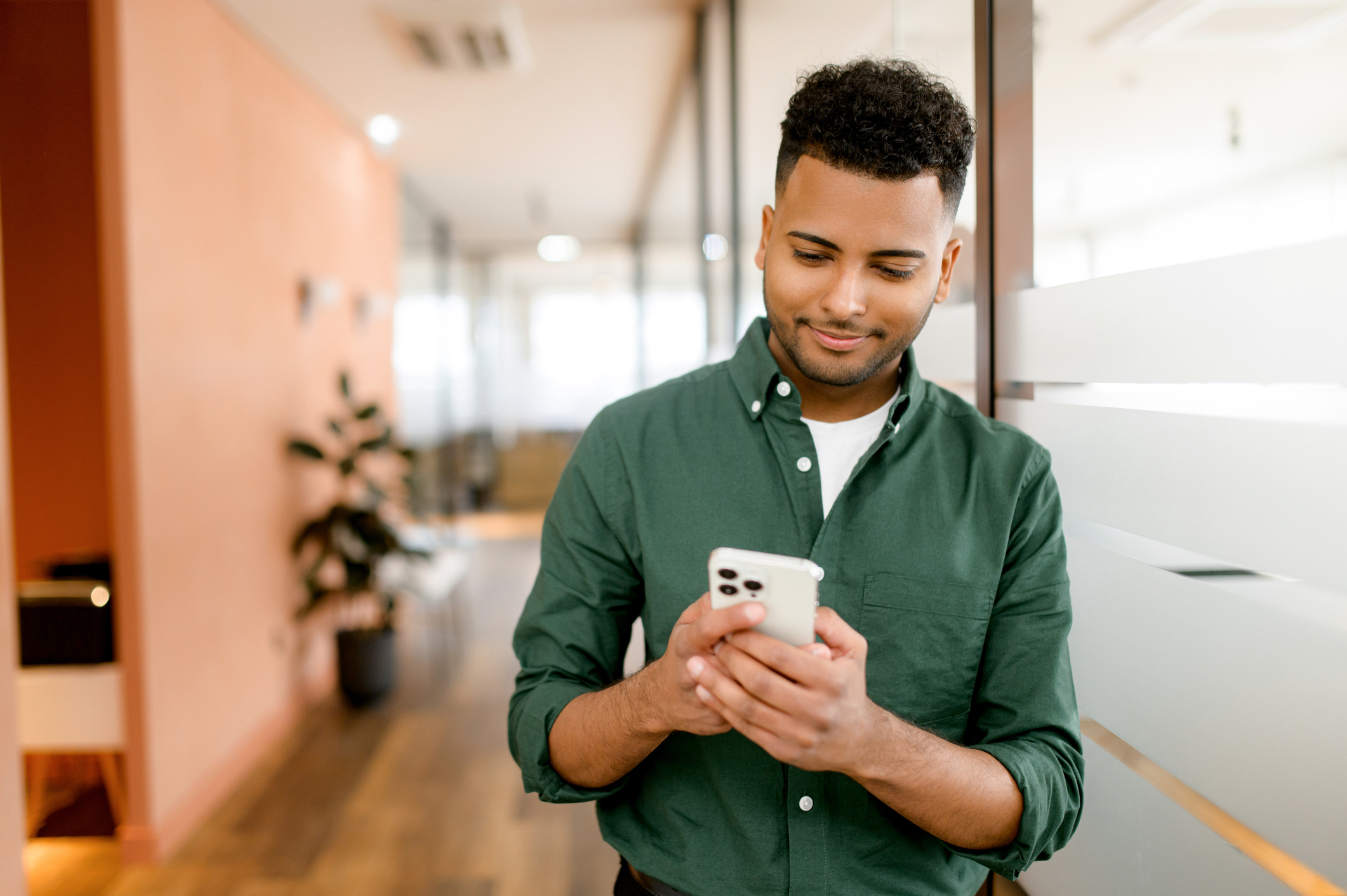 A real estate agent in a green shirt smiles while using his smartphone, standing in a modern office. This image illustrates the convenience of centralizing client communications on platforms like CryptoPay, enabling real estate agents to efficiently manage contacts and streamline interactions.